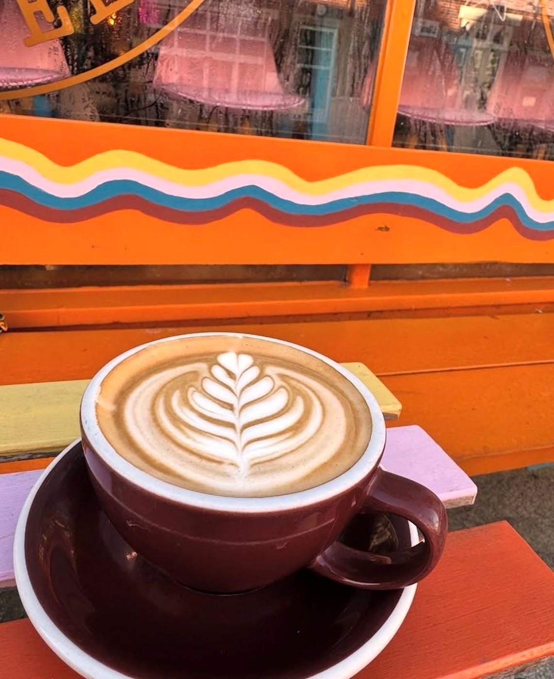 A latte with a leaf in the foam, in a deep red cup, on a bench painted with wavy orange, yellow and blue stripes